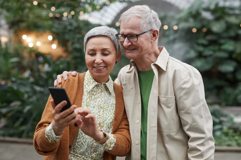 An older couple stands close together outdoors, smiling as she shows him something on her phone.