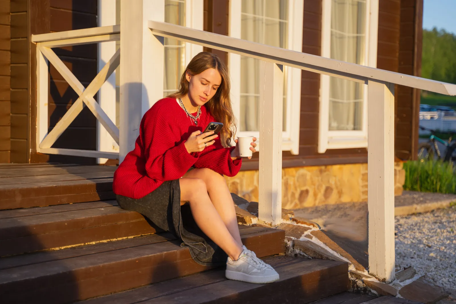 A young woman sits on the wooden front steps of a modest home at golden hour, coffee in one hand, checking her phone.