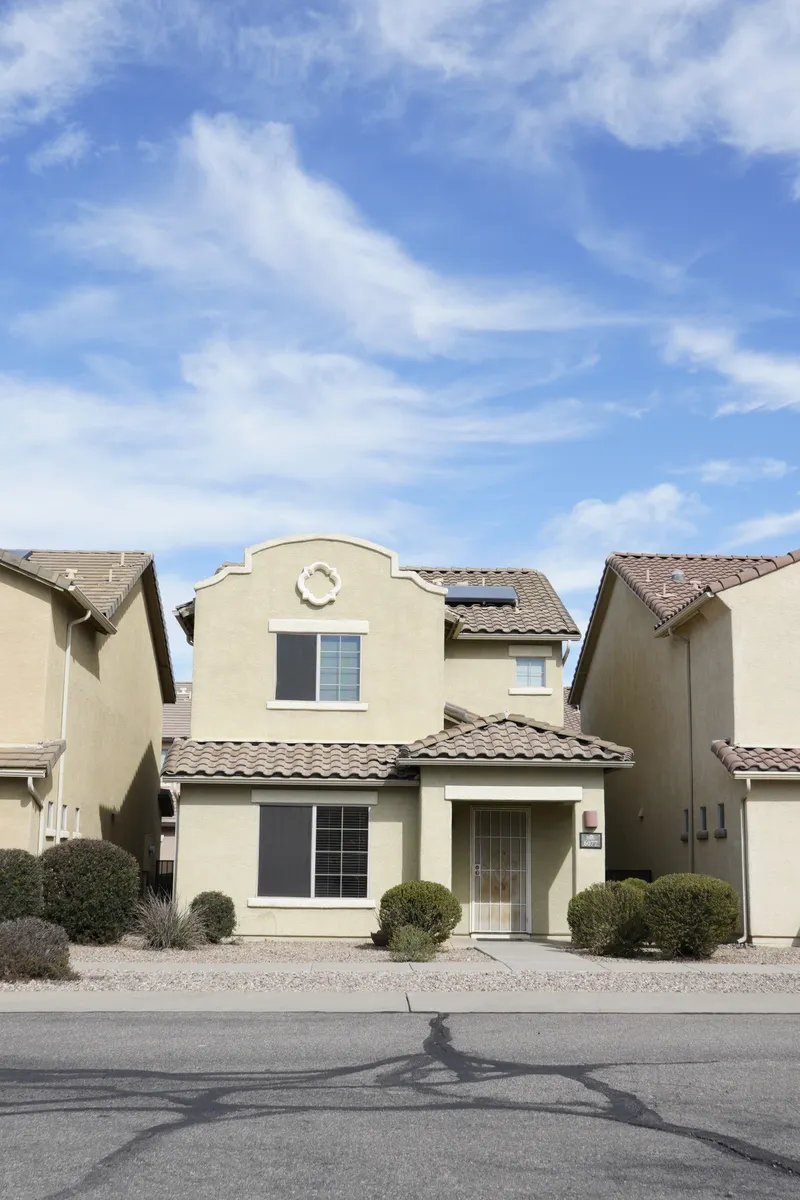 A two-story tan stucco house with a clay tile roof and low desert shrubs under a bright, cloud-streaked sky.