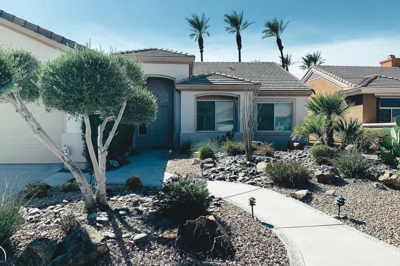 A single-story stucco home with a tile roof, rock-and-cactus desert landscaping, and tall palm trees against a blue sky.