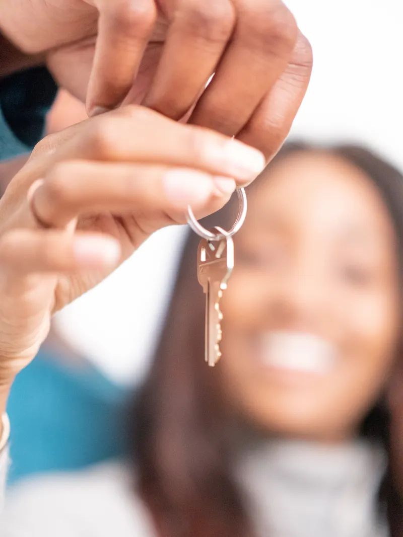 A hand holds up a single house key, with a smiling woman blurred in the background.